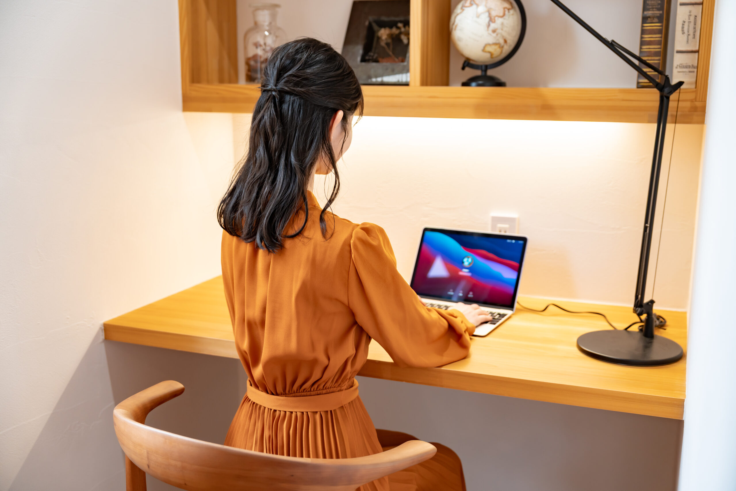 オレンジ色のワンピースを着た女性が、デスクでノートパソコンに向かっている後ろ姿